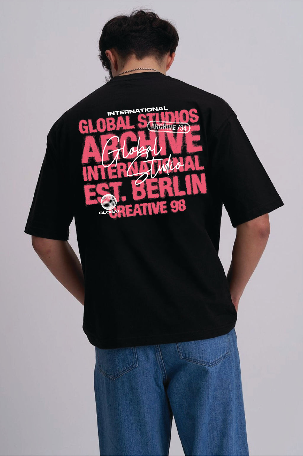 A man viewed from behind wearing a black graphic t-shirt with a large pink and white text print on the back, paired with blue jeans, against a light grey studio background.