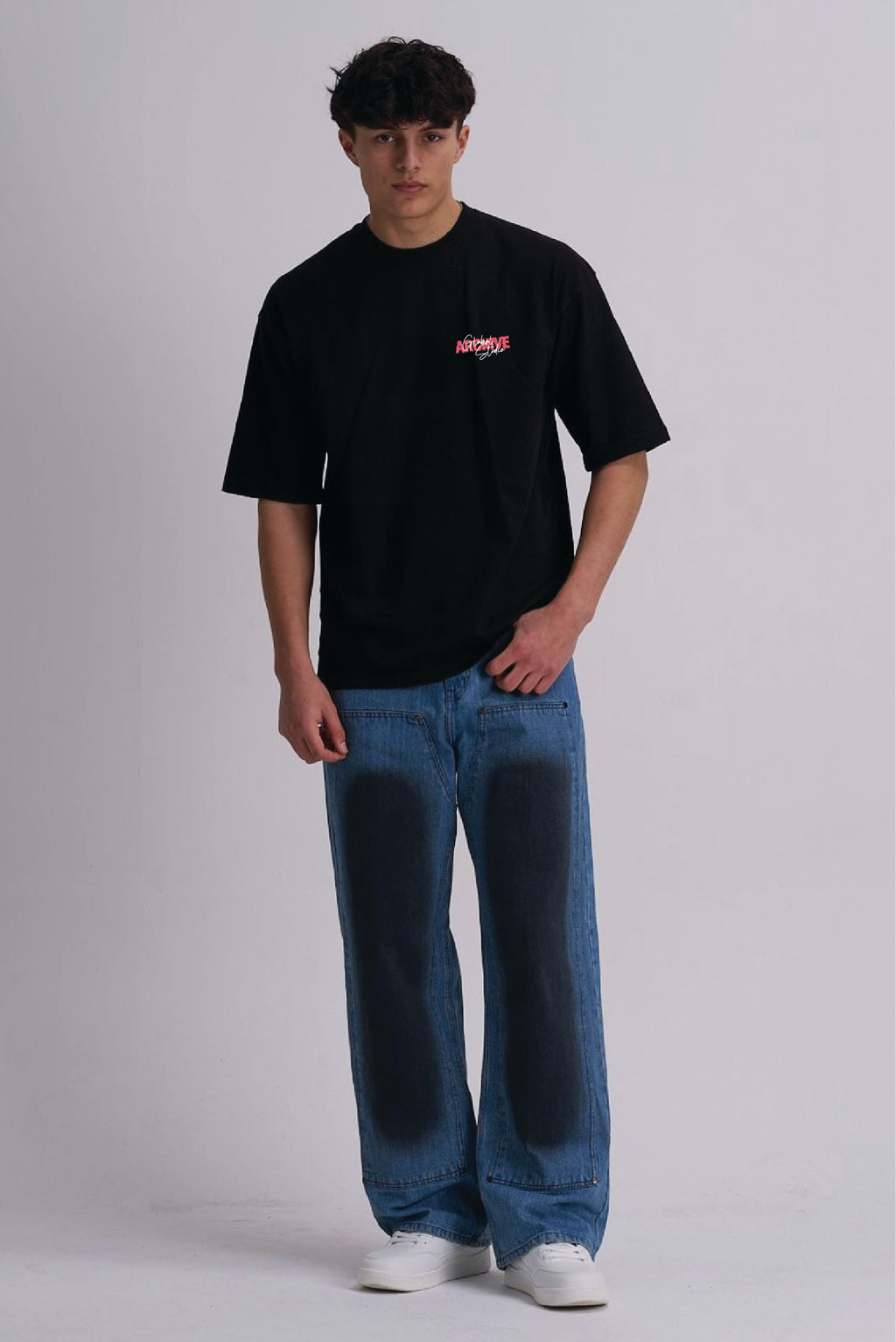 A man modelling a black short-sleeve t-shirt with a small pink and white graphic on the left chest, paired with wide-leg blue jeans featuring dark patches and white sneakers, against a light grey studio background.