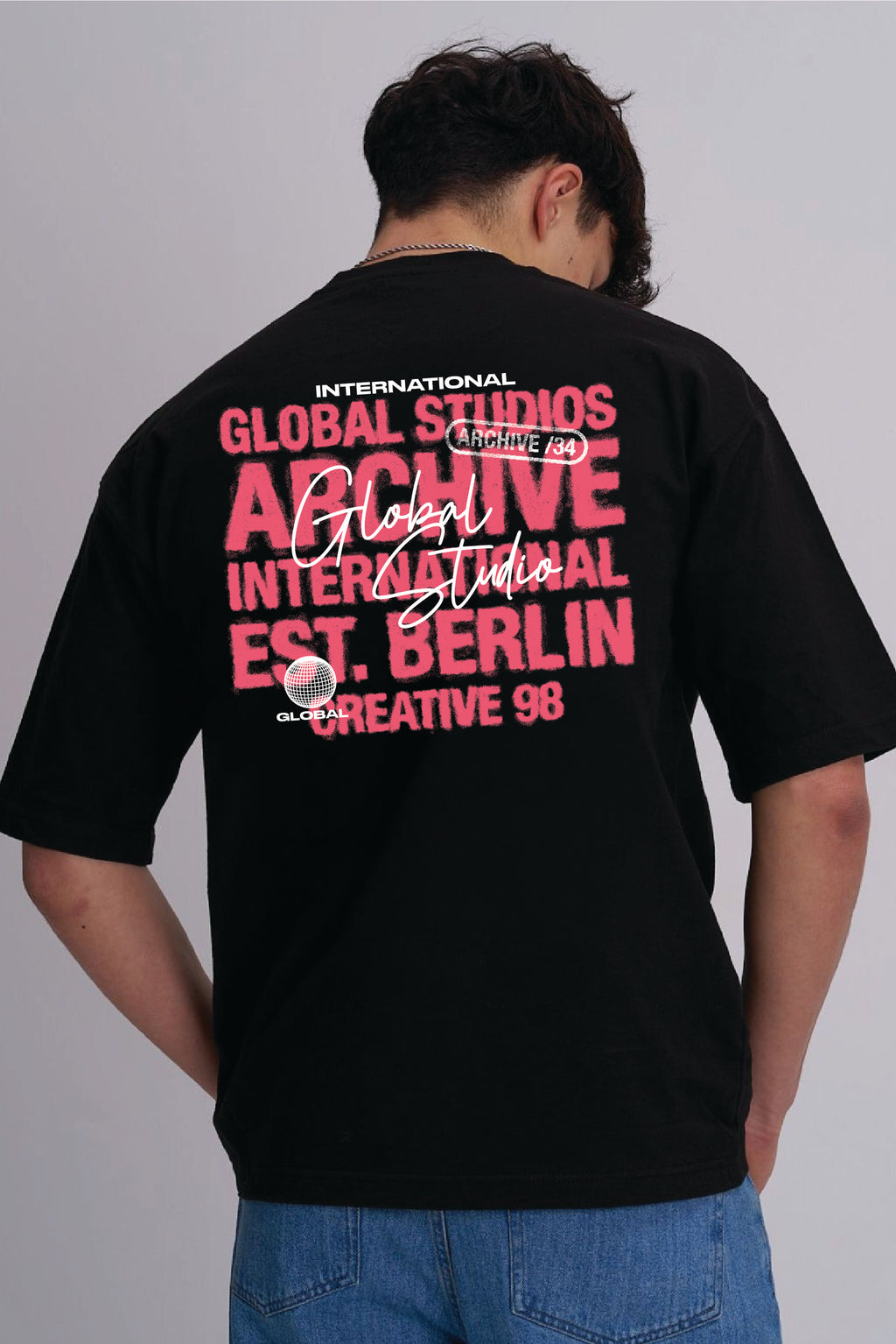 A man viewed from behind wearing a black t-shirt with a large pink and white typography print on the back, paired with blue jeans, against a plain grey studio background.