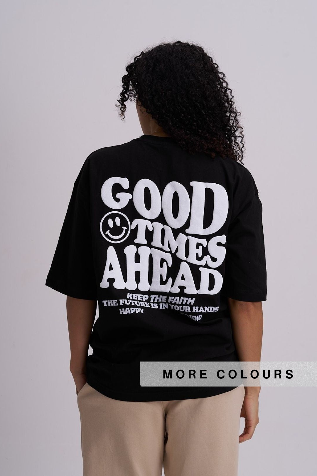A person shown from the back wearing a black oversized T-shirt with large white text reading 'GOOD TIMES AHEAD' and smaller text below, paired with light beige pants. A small smiley face is integrated into the design.