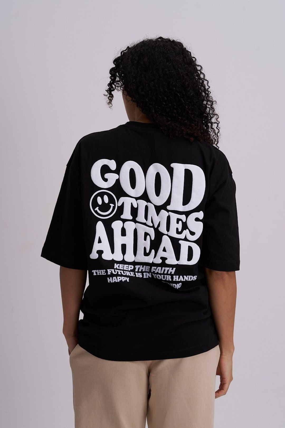 Female wearing a black t-shirt with 'Good Times Ahead' text and smiley face on the back, standing against a plain background.