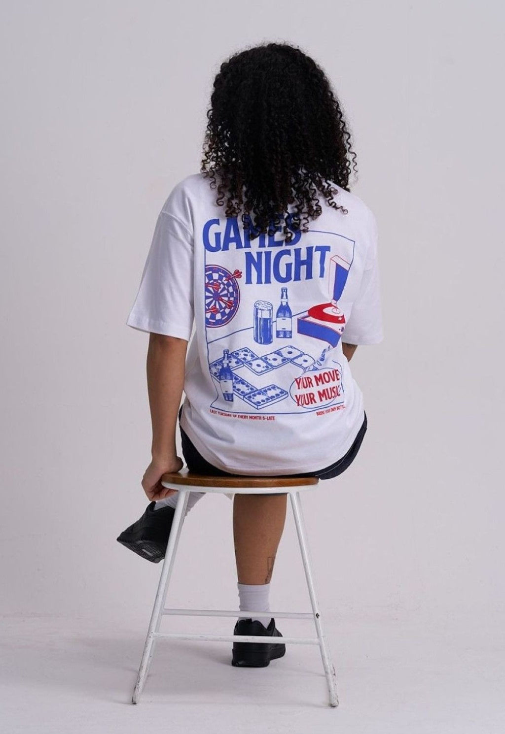 Female wearing a white t-shirt with 'Game Night' graphics sitting on a stool against a plain background