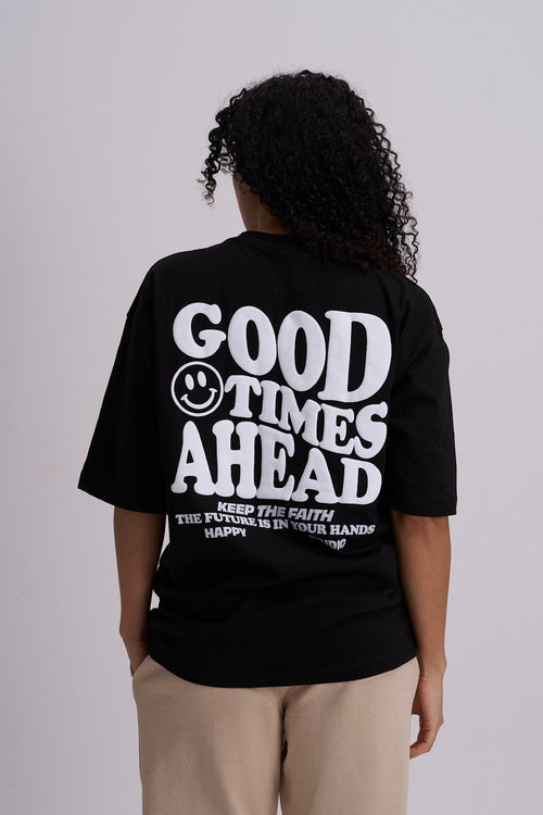  Female wearing a black t-shirt with 'Good Times Ahead' text and smiley face on the back, standing against a plain background.