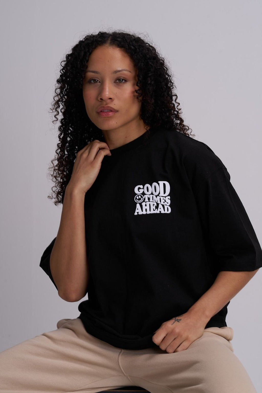 Woman wearing a black t-shirt with 'Good Times Ahead' text on a plain background, sat on a stool
