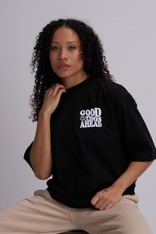 Woman wearing a black t-shirt with 'Good Times Ahead' text on a plain background, sat on a stool