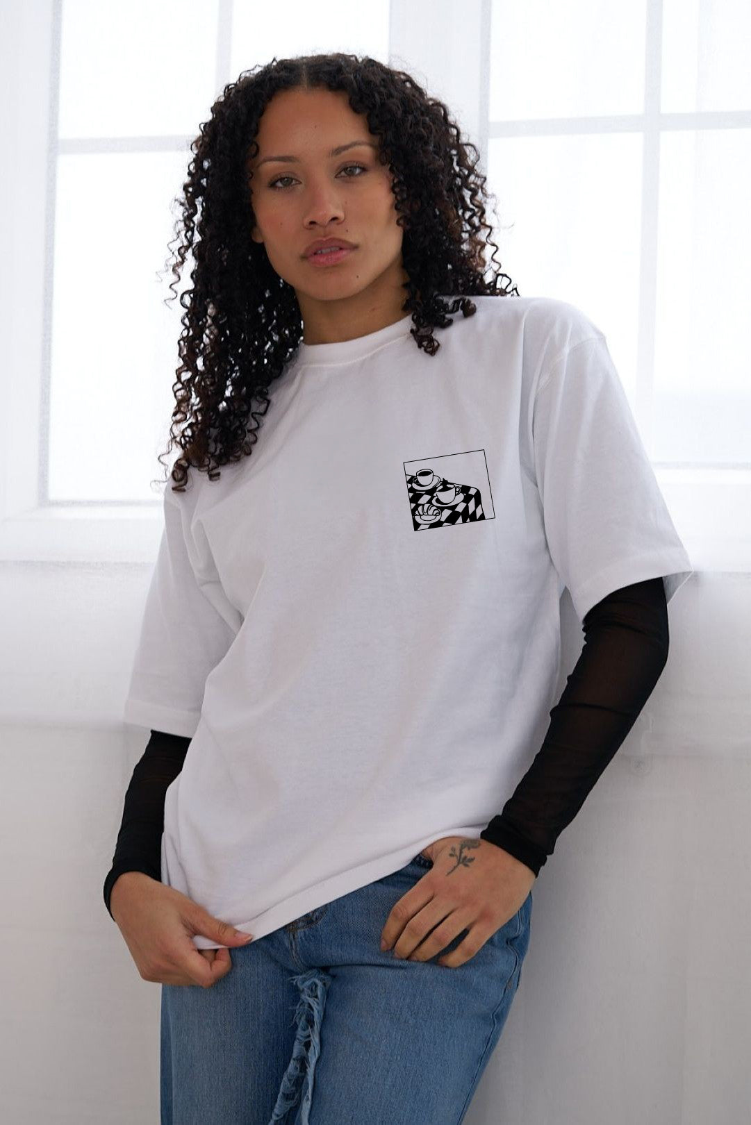 A woman wearing a white oversized T-shirt with a small black coffee cup graphic on the left chest, layered over a black long-sleeve mesh top and paired with blue jeans, standing in front of a bright window.