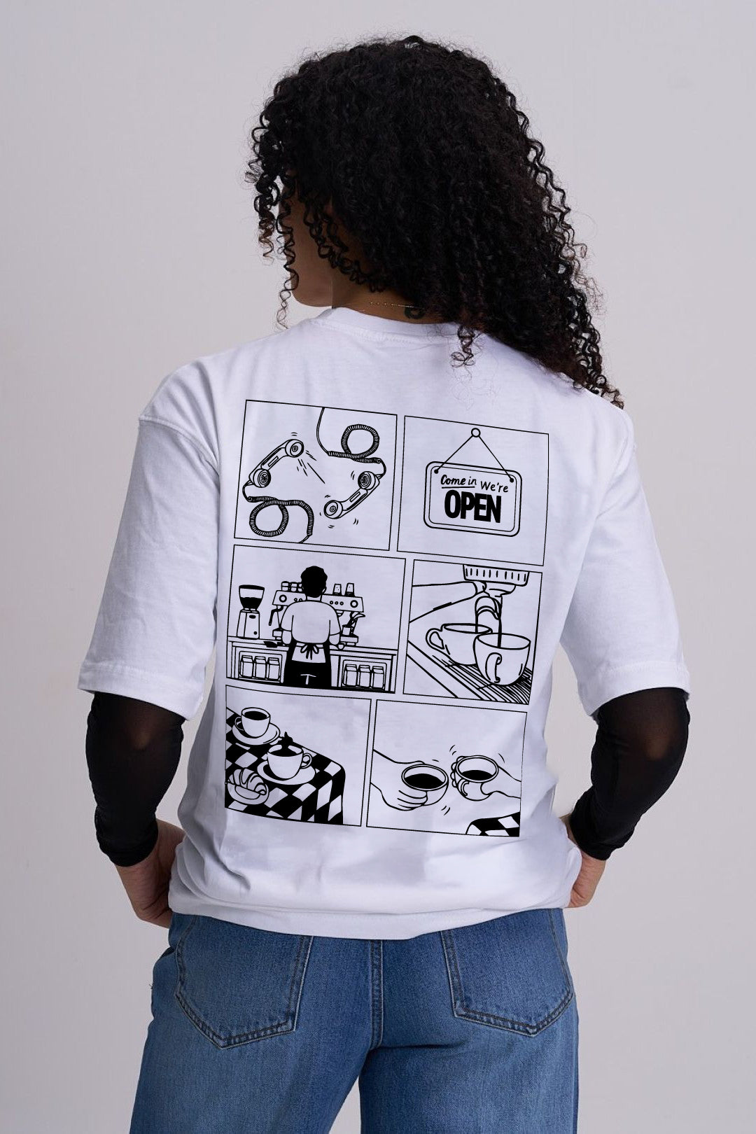 A female shown from the back wearing a white graphic T-shirt with a black comic-style coffee print and blue jeans, photographed against a light grey studio background.