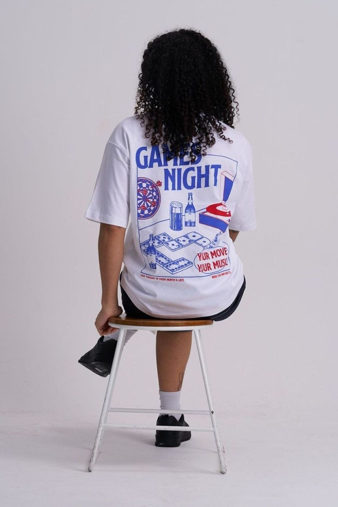 Woman wearing a white t-shirt with 'Game Night' graphics sitting on a stool against a plain background