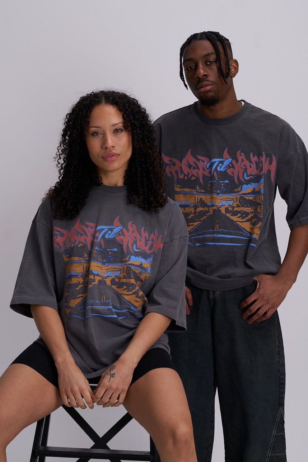 Male and female wearing matching desert highway graphic t-shirts against a plain background