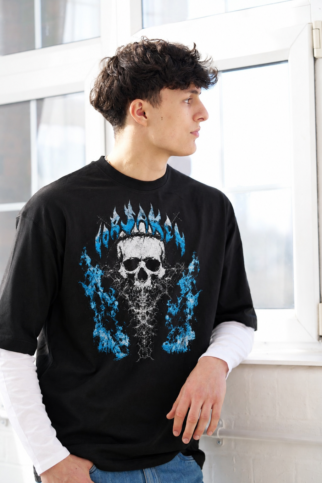 A man wearing a black long-sleeve t-shirt with a large graphic of a skull surrounded by blue flames and the word 'FORSAKEN' in a flame-like font, layered over a white long-sleeve shirt and blue jeans.