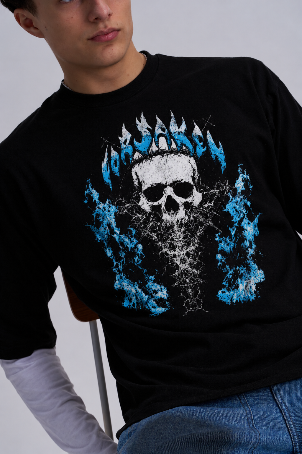A man wearing a black long-sleeve t-shirt with a graphic print of a skull wearing a crown of blue flames, layered over a white long-sleeve shirt and blue jeans.