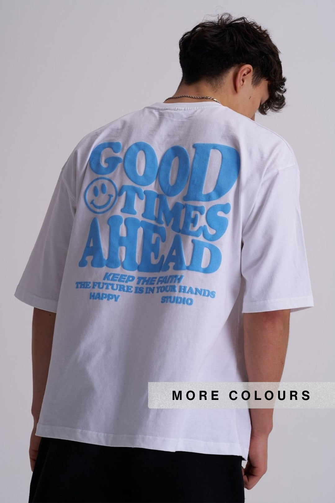 A man viewed from behind wearing a white oversized T-shirt with a large blue graphic print reading 'GOOD TIMES AHEAD' and smaller text below, against a light grey studio background. 