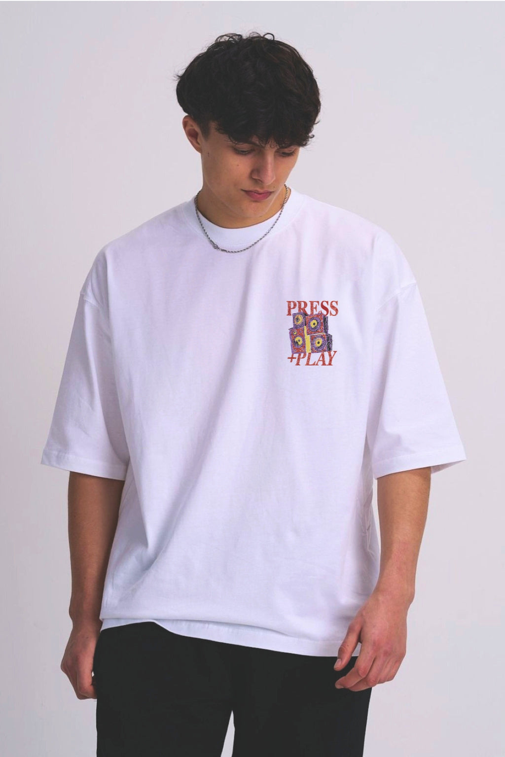 A man wearing an oversized white t-shirt with a multicolour music speaker graphic on the left chest that reads 'PRESS PLAY'.