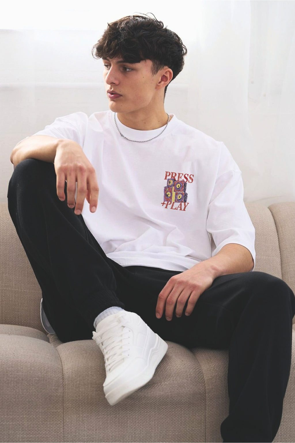 A young man sitting on a beige couch wearing a white graphic t-shirt with an illustrated speaker graphic and 'PRESS PLAY' printed on the chest, black sweatpants, and white sneakers.