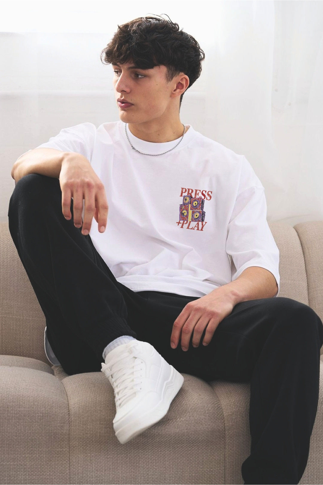 A young man sitting on a beige couch wearing a white graphic t-shirt with an illustrated speaker graphic and 'PRESS PLAY' printed on the chest, black sweatpants, and white sneakers.