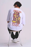 A male seated on a metal stool, viewed from the back, wearing a white oversized T-shirt with a large multicolour music speaker graphic print reading “PRESS PLAY” on the back