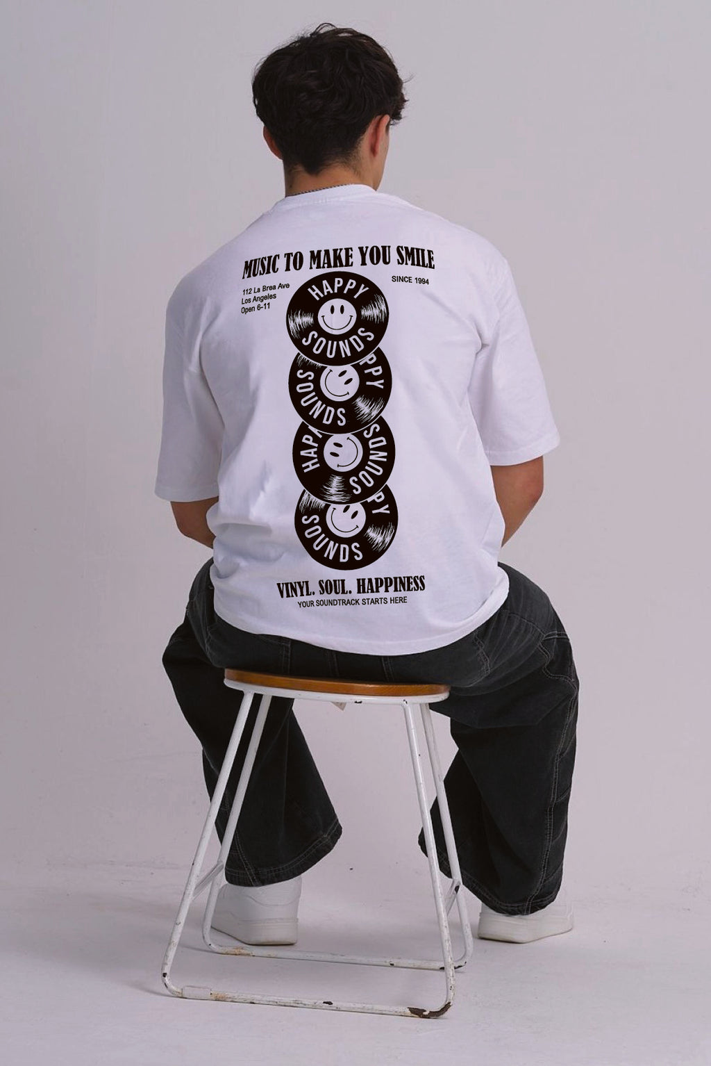 A man sitting on a metal stool, viewed from behind, wearing a white graphic t-shirt with a black vinyl record smiley face design on the back.
