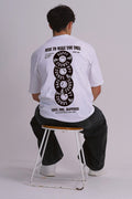 A man sitting on a metal stool, viewed from behind, wearing a white graphic t-shirt with a black vinyl record smiley face design on the back.