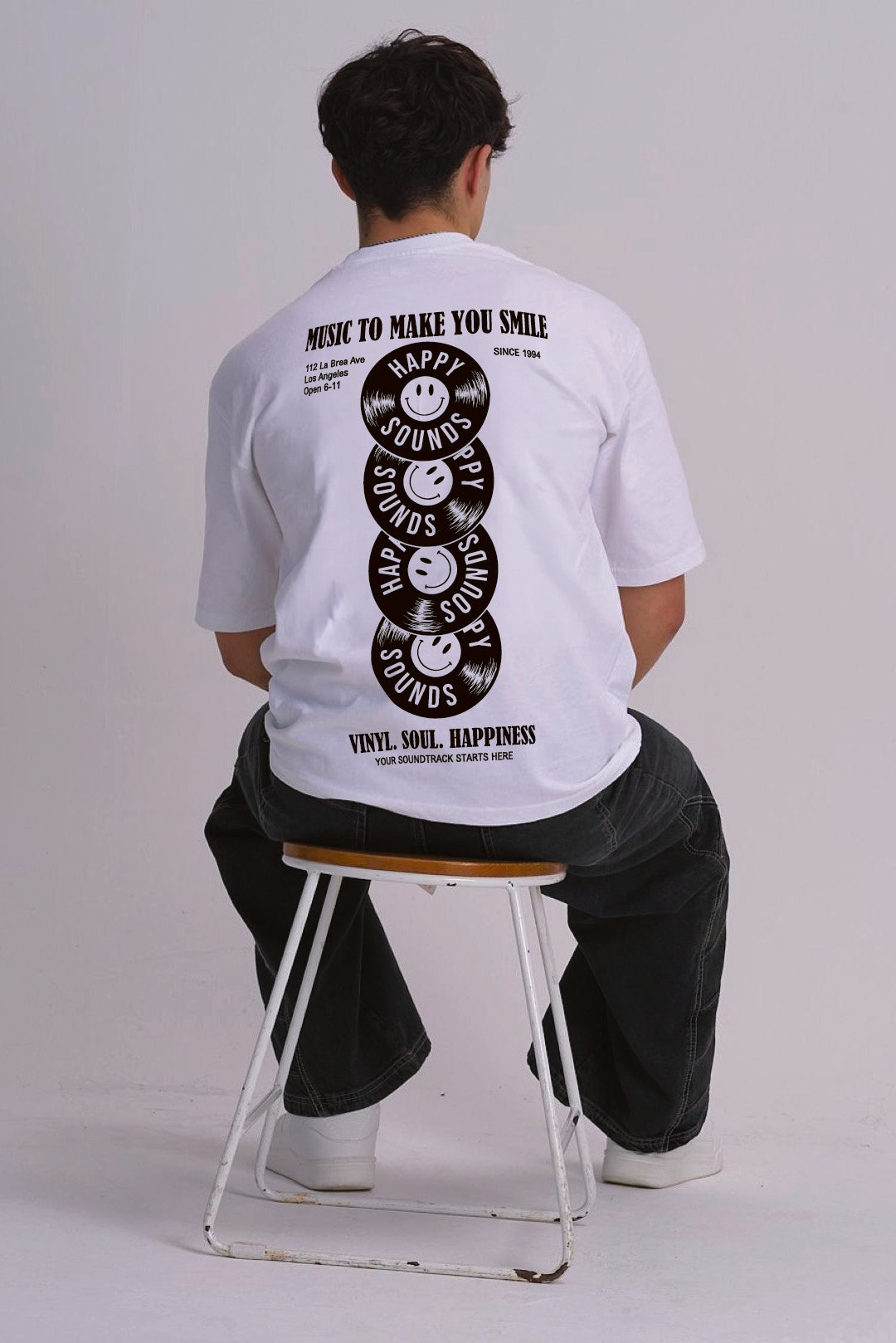 A man sitting on a metal stool, viewed from behind, wearing a white graphic t-shirt with a black vinyl record smiley face design on the back.