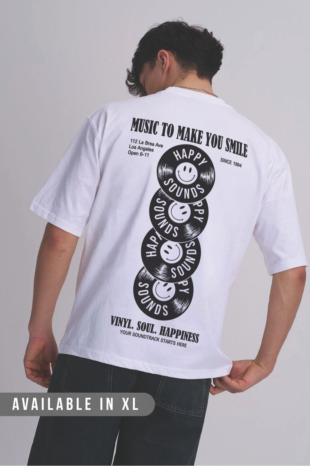 A man wearing a white oversized t-shirt featuring a black graphic back print of stacked vinyl records with smiley faces and text that reads 'MUSIC TO MAKE YOU SMILE' and 'HAPPY SOUNDS'