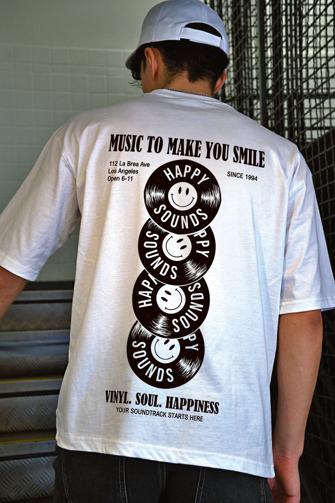 A man viewed from behind wearing a white t-shirt with a large black graphic print on the back. The graphic features stacked vinyl records with smiley faces and the text 'HAPPY SOUNDS' and 'MUSIC TO MAKE YOU SMILE'.