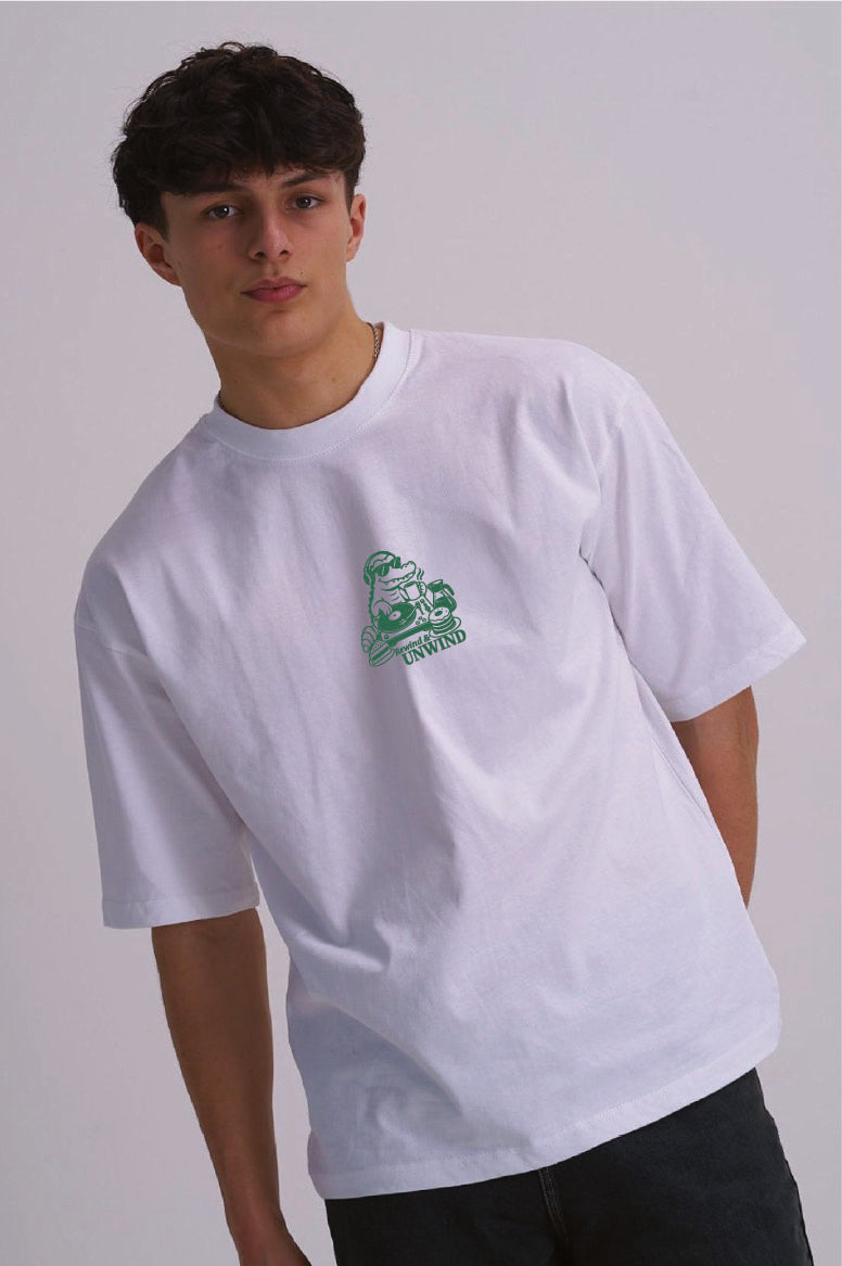 A young man wearing a white crewneck t-shirt with a green cartoon mascot graphic on the centre chest, paired with dark jeans, against a plain light grey background.