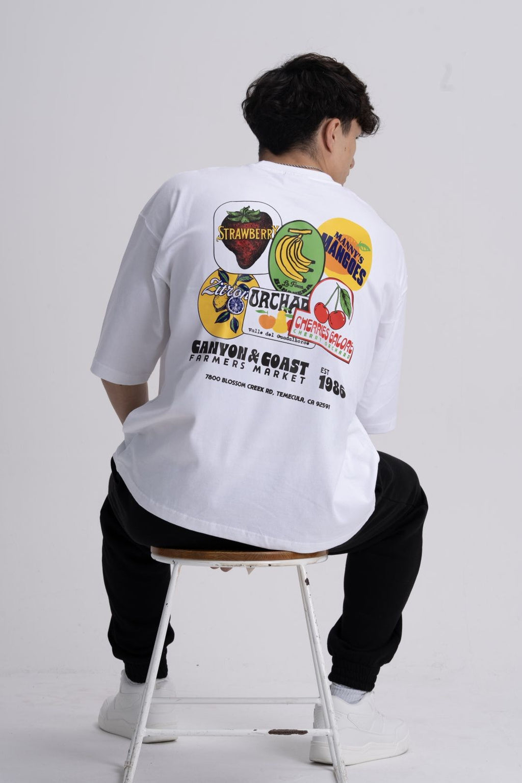 male wearing a white t-shirt with colourful food graphics on the back, sitting on a stool against a plain background.
