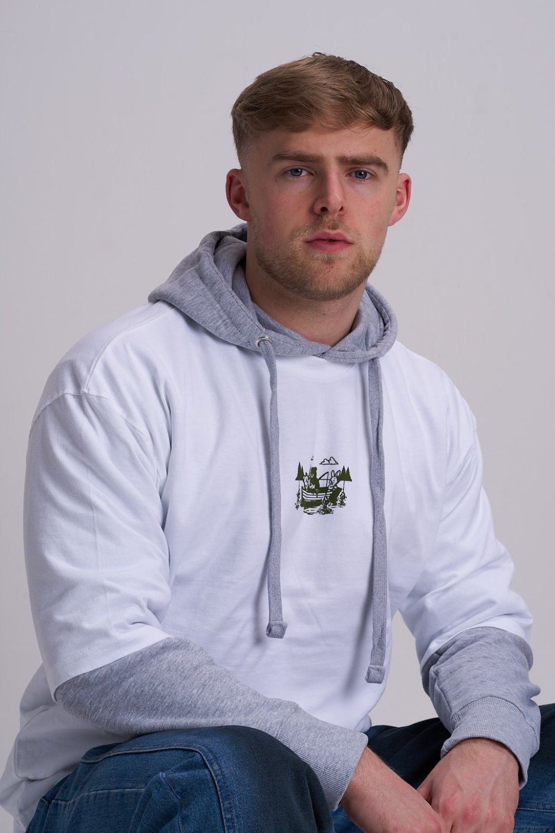Man wearing a grey  hoodie and white tee with a fishing boat logo on a plain background