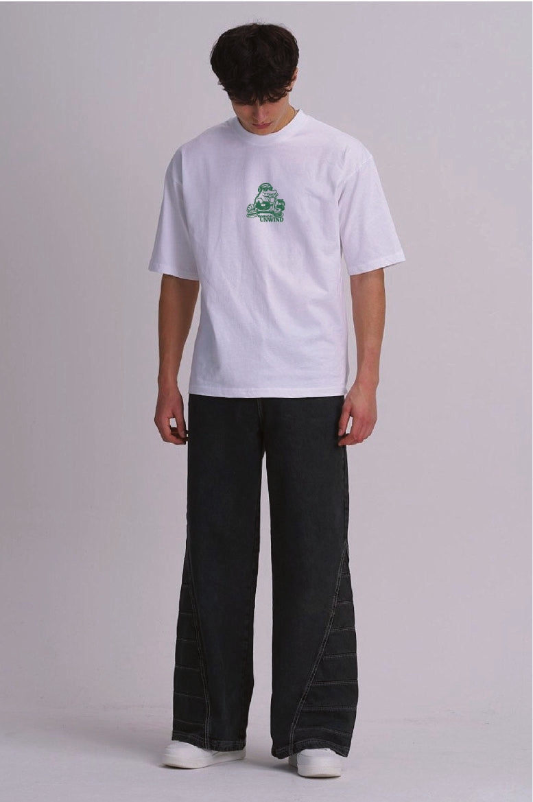 A model wearing a white short-sleeve t-shirt with a small green graphic on the chest, paired with dark wide-leg jeans and white sneakers, standing against a light grey studio background.