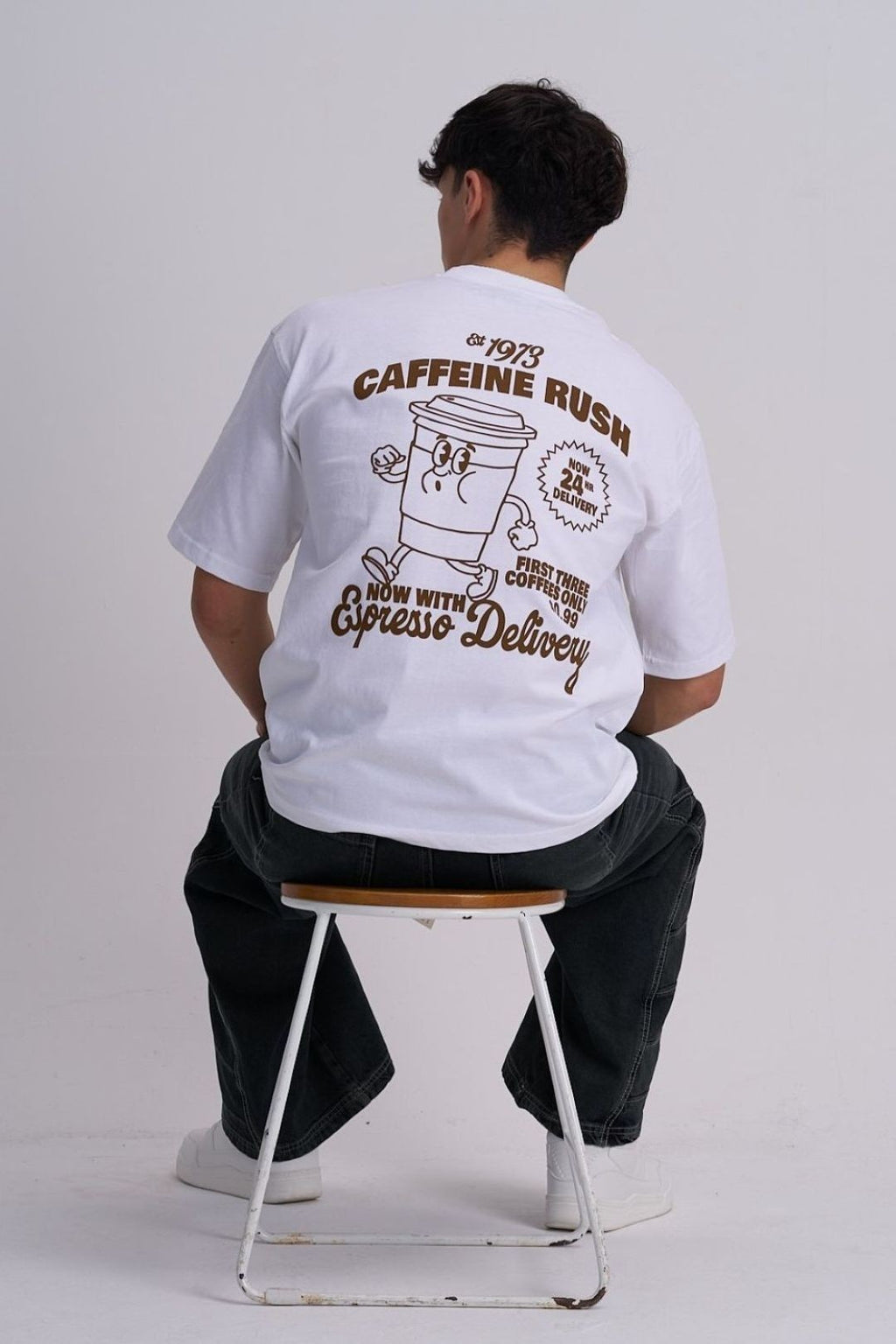 Man wearing a white t-shirt with coffee-themed graphics and text, sitting on a stool.