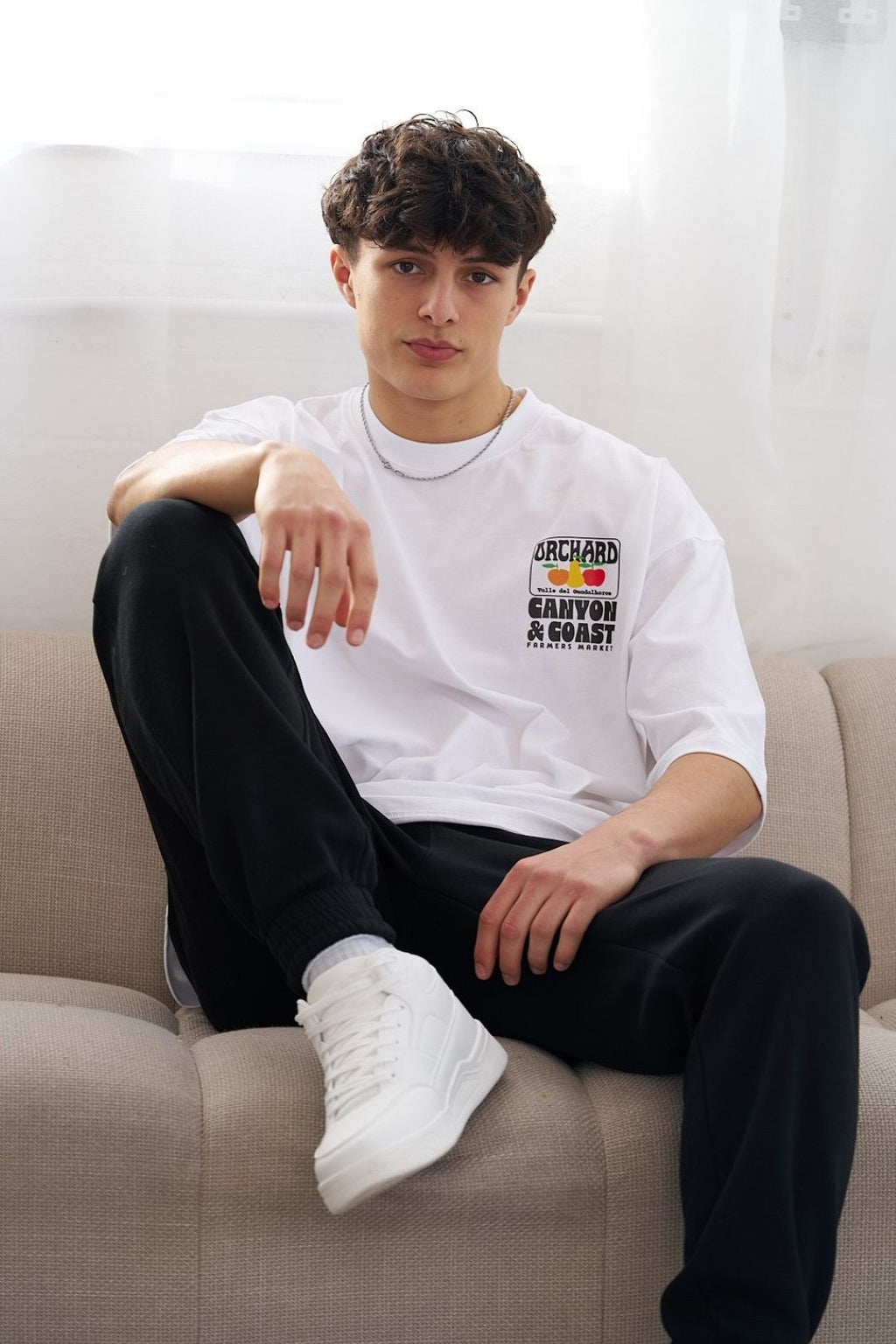Man wearing a white t-shirt with a colourful logo sitting on a beige couch.