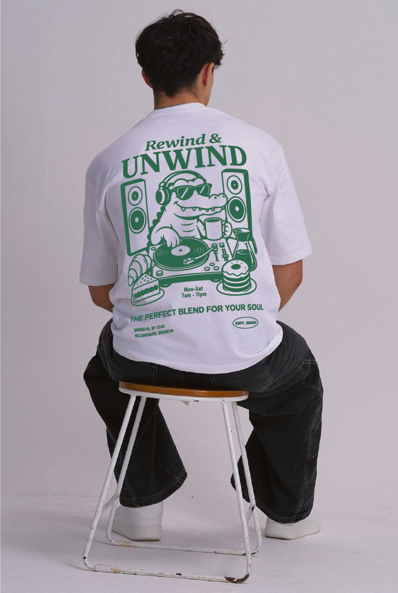 A man sitting on a stool, viewed from behind, wearing a white t-shirt with a large green graphic print on the back. The graphic features the text 'Rewind & UNWIND' and a crocodile DJ illustration.