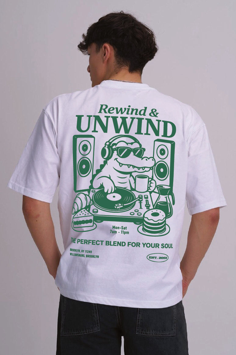A man viewed from behind wearing a white t-shirt with a large green graphic print on the back. The graphic features a crocodile DJ with headphones, speakers, and vinyl records, with the text 'Rewind & UNWIND' and 'THE PERFECT BLEND FOR YOUR SOUL'.