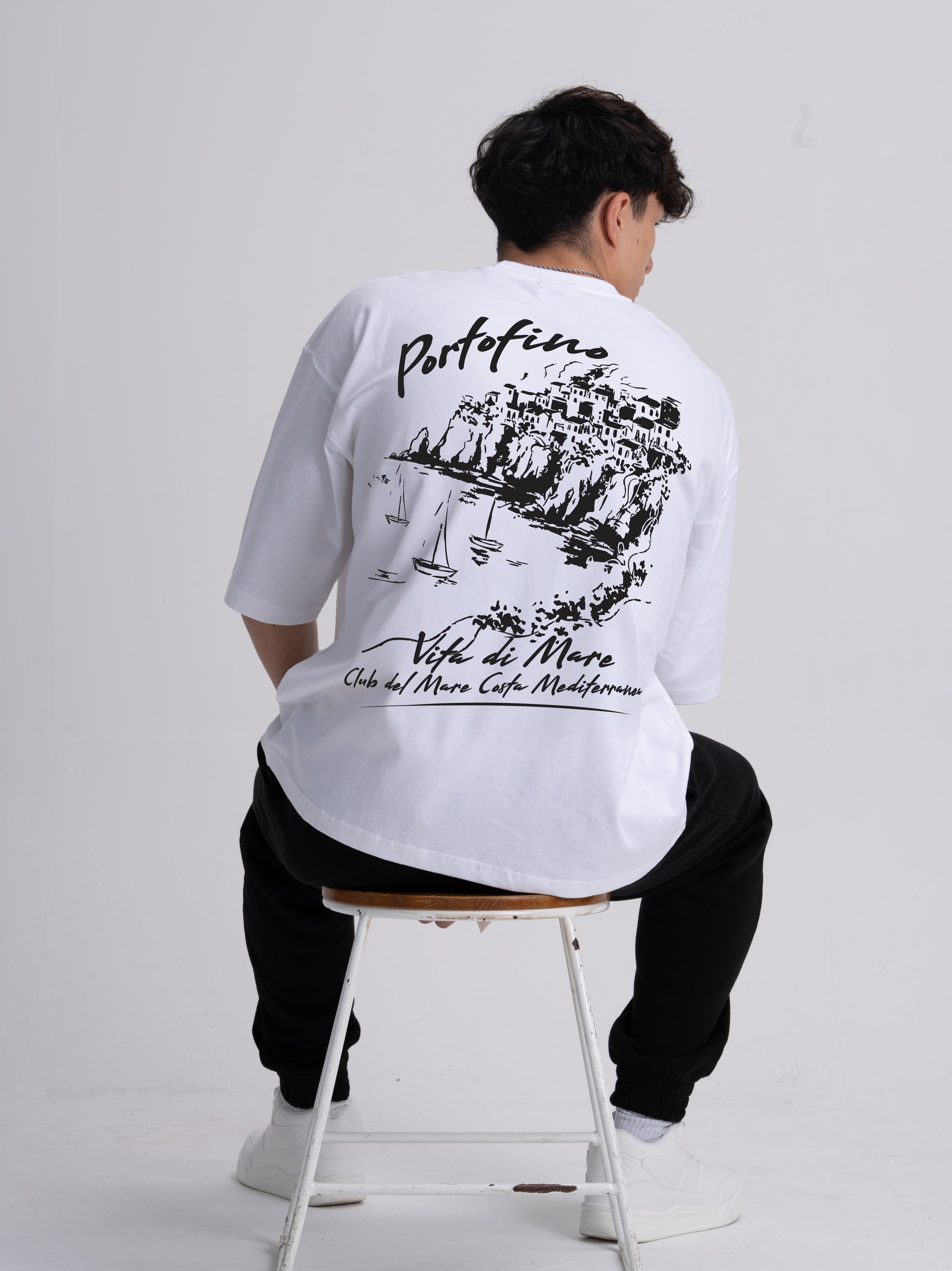 Person wearing a white t-shirt with black coastal line drawing graphics and text, sitting on a stool against a plain background