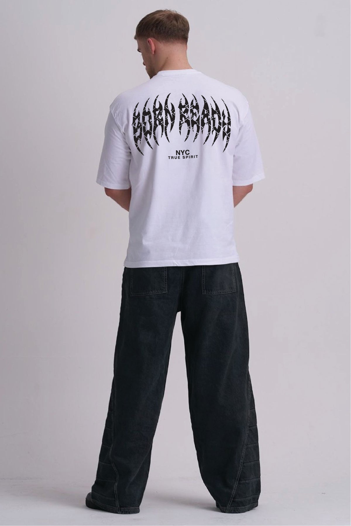 A man viewed from behind wearing a white t-shirt with a large black 'BORN READY' graphic on the back, paired with dark wide-leg jeans, against a light grey studio background.