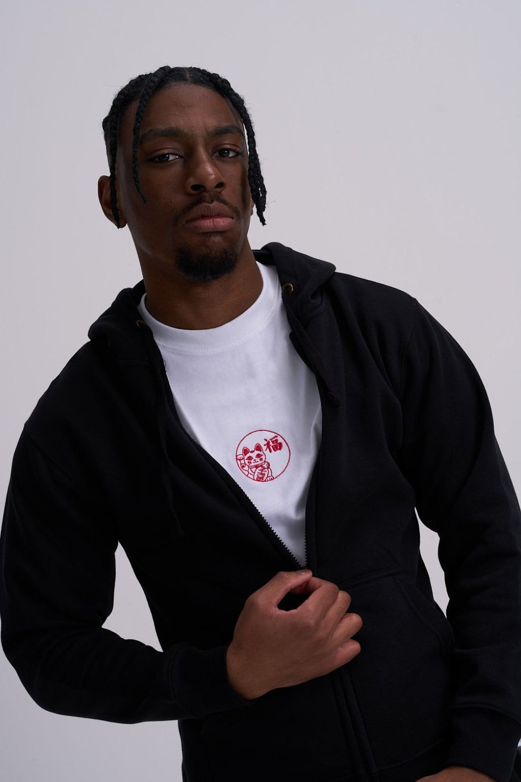 Man wearing a black hoodie over a white shirt with a red lucky cat embroidery on a plain background