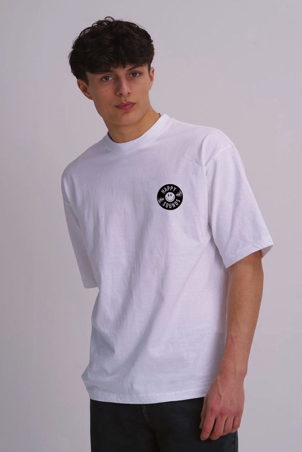 A male wearing a white short-sleeve crewneck t-shirt with a black vinyl record chest graphic, paired with dark jeans, photographed against a neutral light grey background.