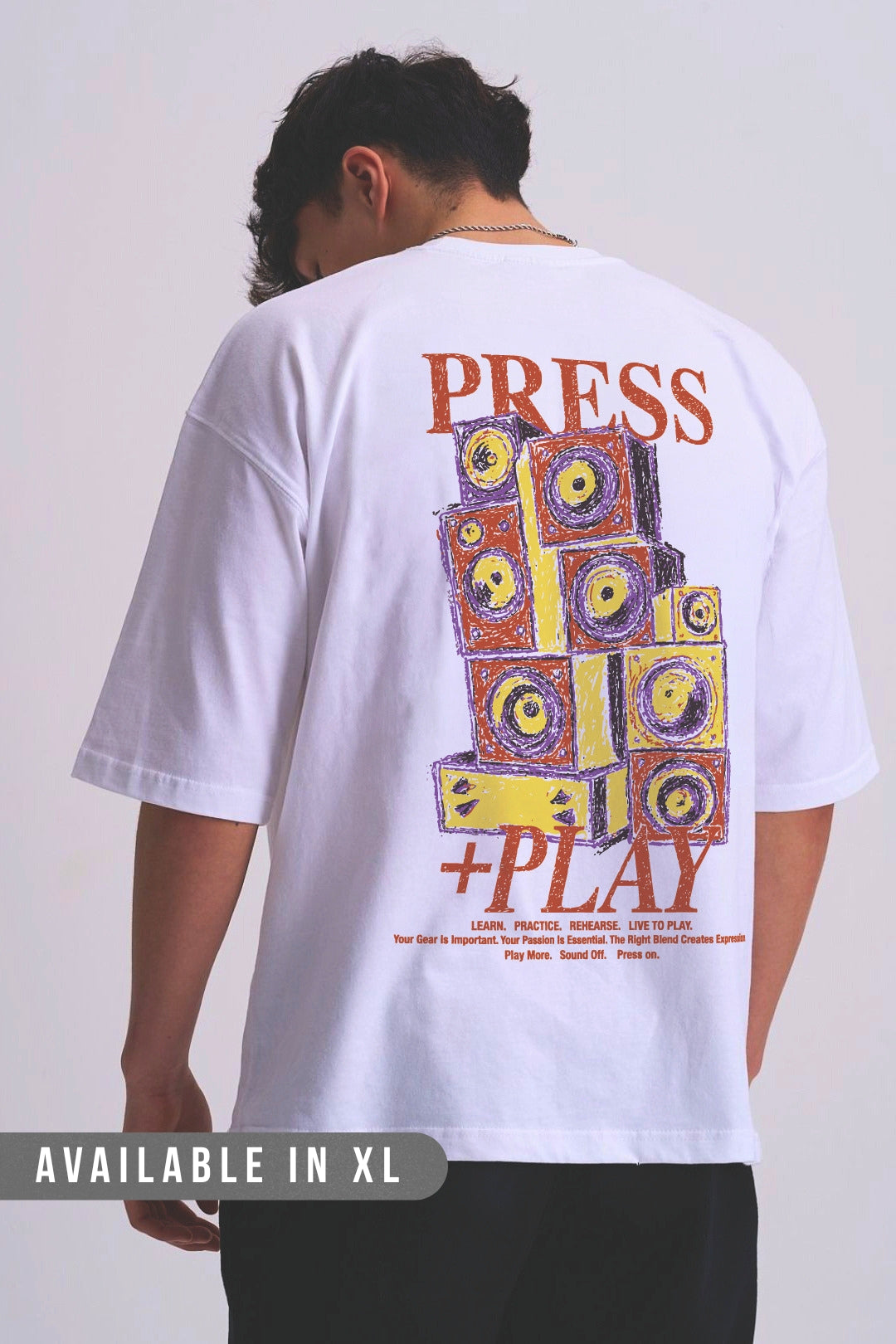 A man viewed from behind wearing a white oversized T-shirt with a large colourful graphic on the back reading 'PRESS +PLAY' and illustrated speaker-like blocks. 