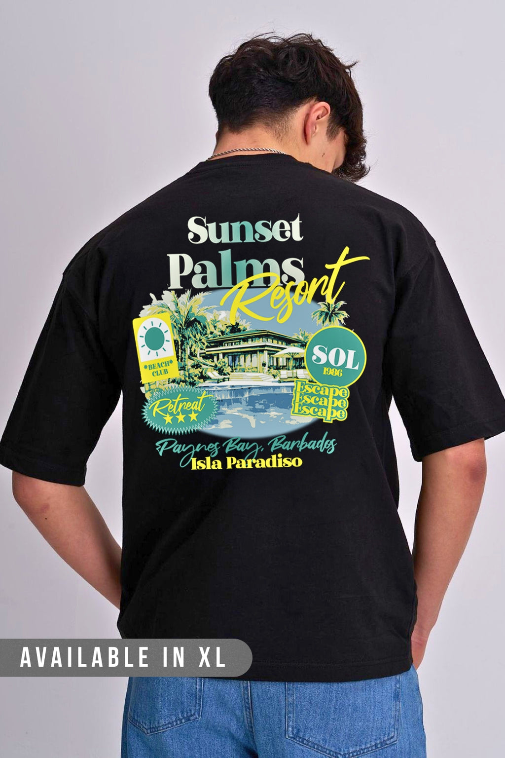 A man shown from the back wearing a black t-shirt with a large colourful graphic print of 'Sunset Palm Resort' on the back, paired with blue jeans against a light grey studio background.