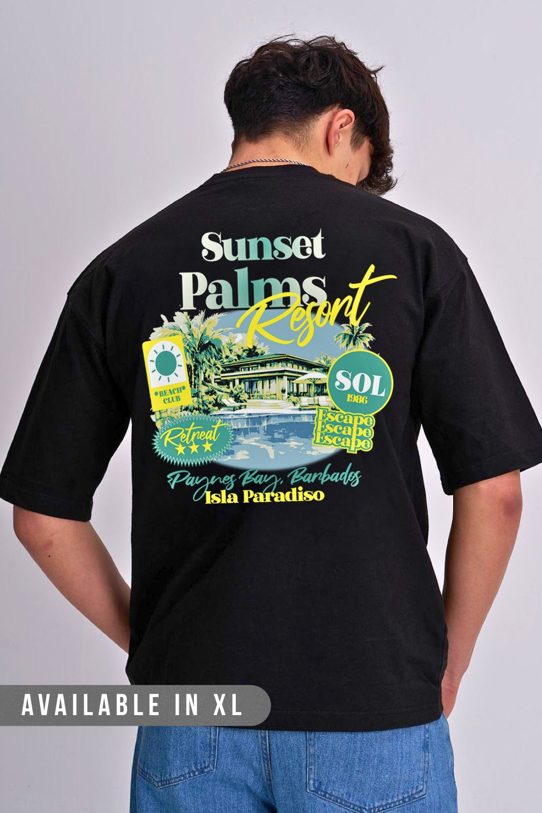 A man shown from the back wearing a black t-shirt with a large colourful graphic print of 'Sunset Palm Resort' on the back, paired with blue jeans against a light grey studio background.
