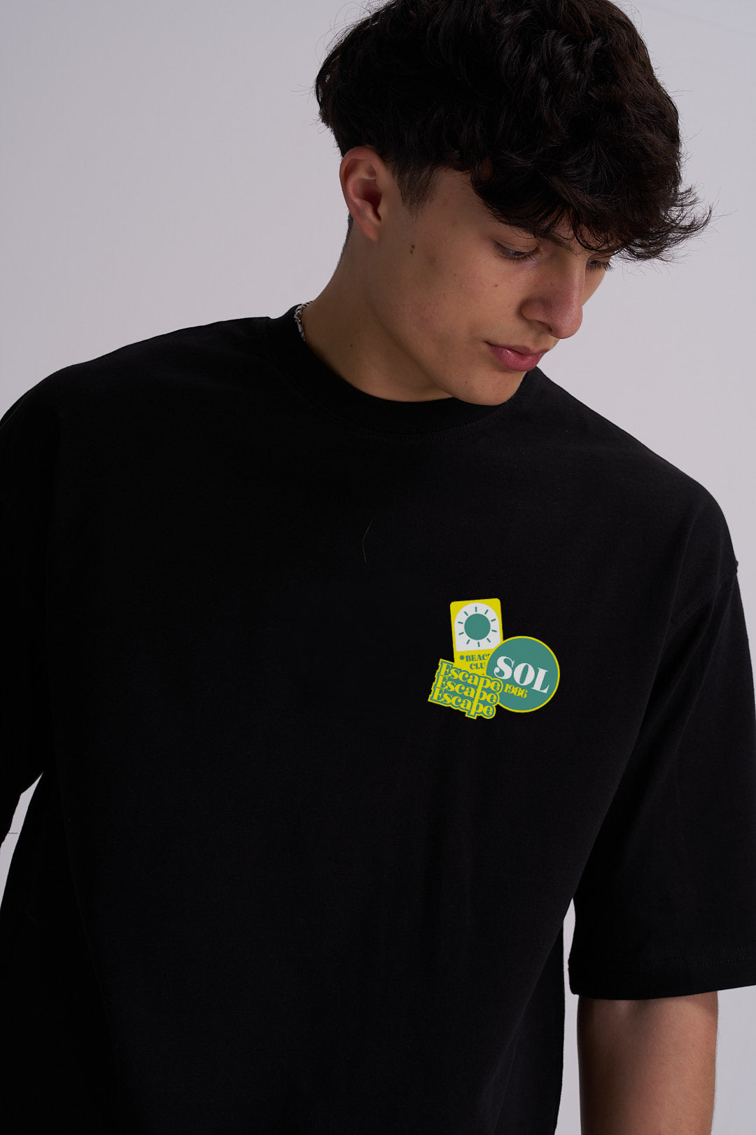 A man wearing a black crewneck t-shirt with a small graphic patch on the left chest featuring a yellow and green design with the text 'SOL'.