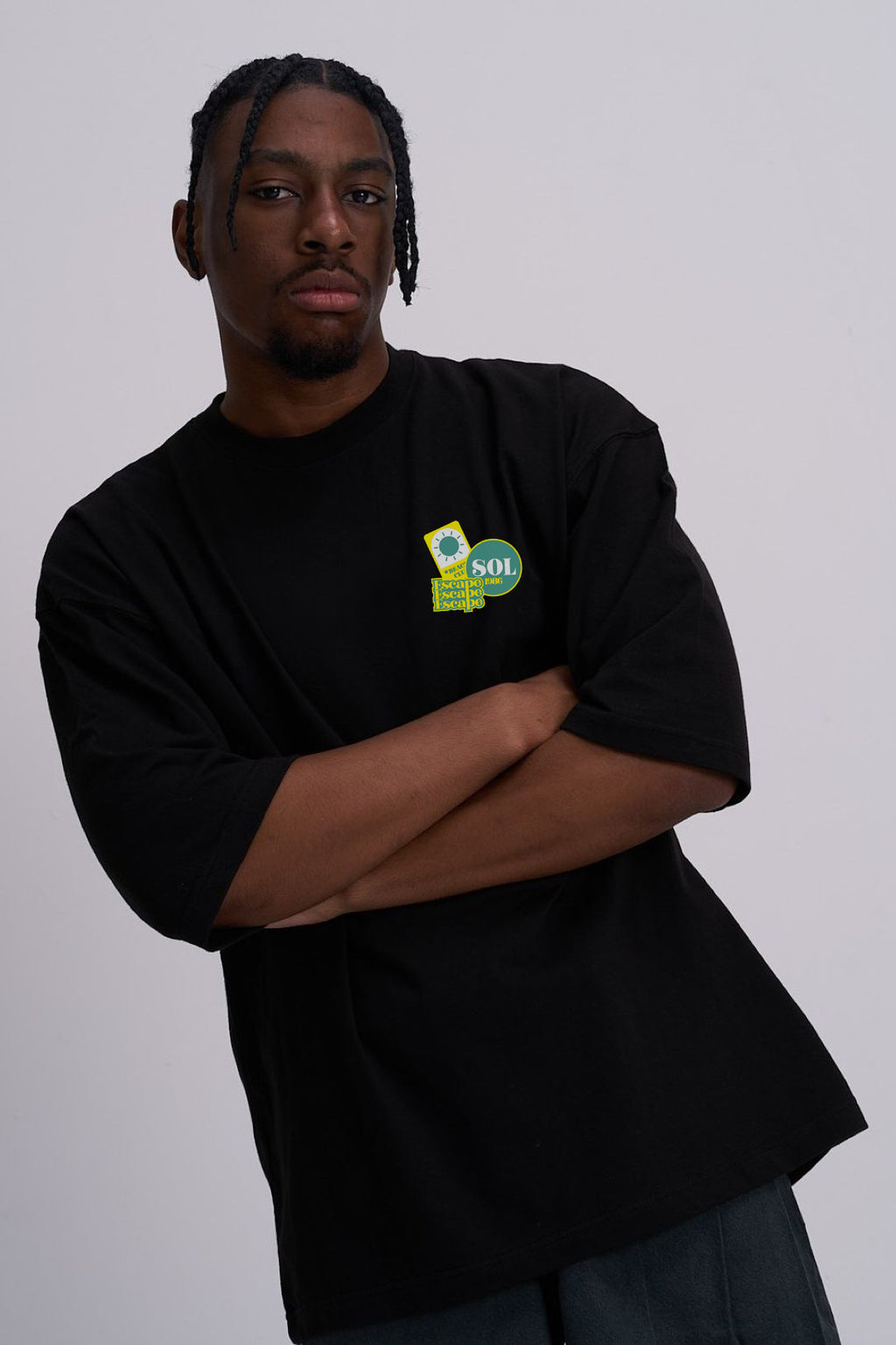 A man wearing a black oversized t-shirt with a small green and yellow graphic on the left chest, posed with arms crossed against a plain light grey background.