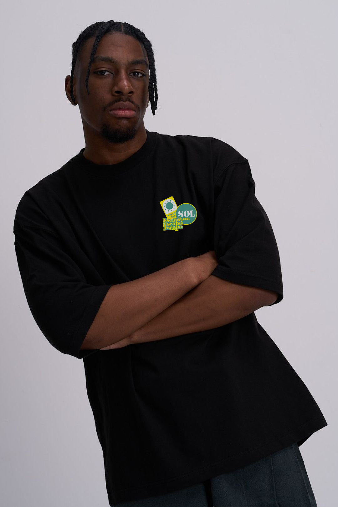 A man wearing a black oversized t-shirt with a small green and yellow graphic on the left chest, posed with arms crossed against a plain light grey background.