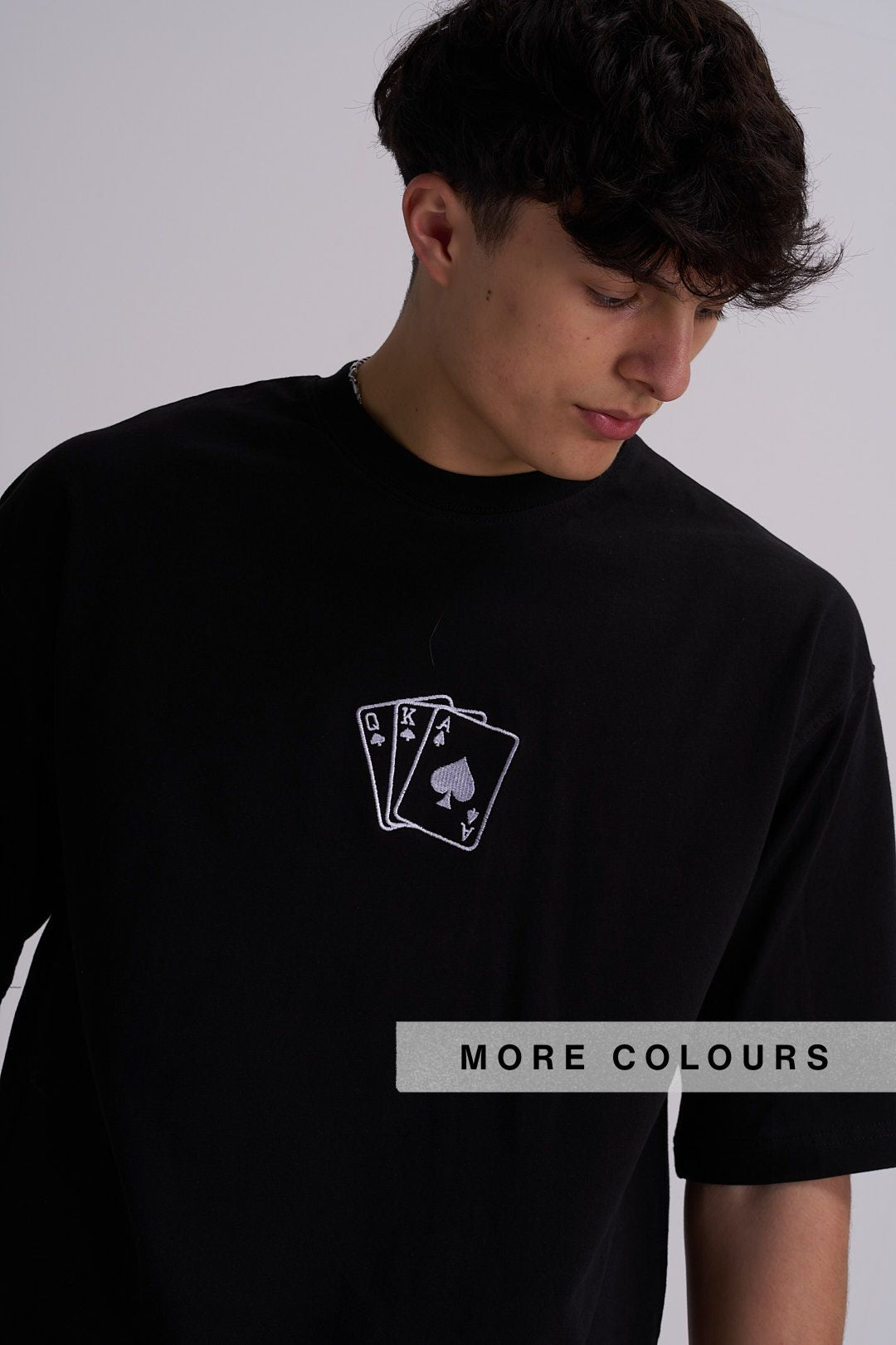A man wearing a black crewneck t-shirt with a white embroidered graphic of three playing cards (King of Spades, Queen of Spades, and Ace of Spades) centre chest. 