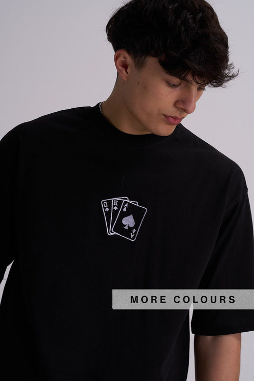 A man wearing a black crewneck t-shirt with a white embroidered graphic of three playing cards (King of Spades, Queen of Spades, and Ace of Spades) centre chest. 