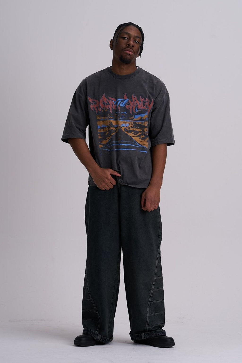 Man wearing a skull highway graphic t-shirt and dark pants on a white background