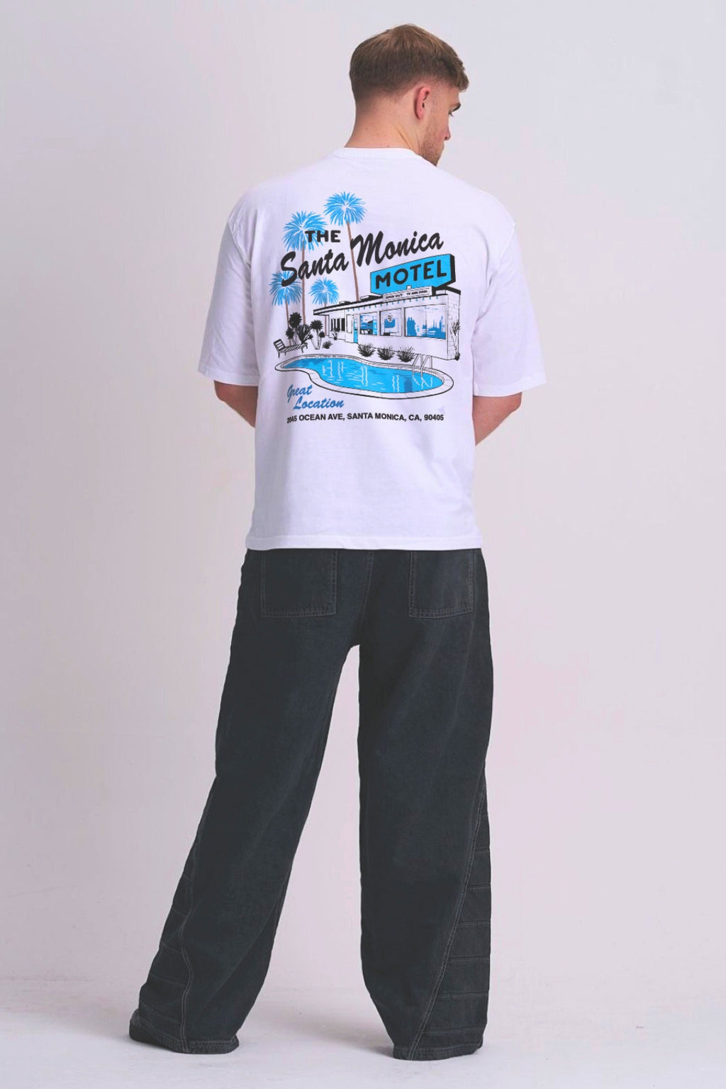 A man viewed from behind wearing a white t-shirt with a blue and black retro 'The Santa Monica Motel' graphic print, paired with dark grey wide-leg jeans, against a light grey studio background.