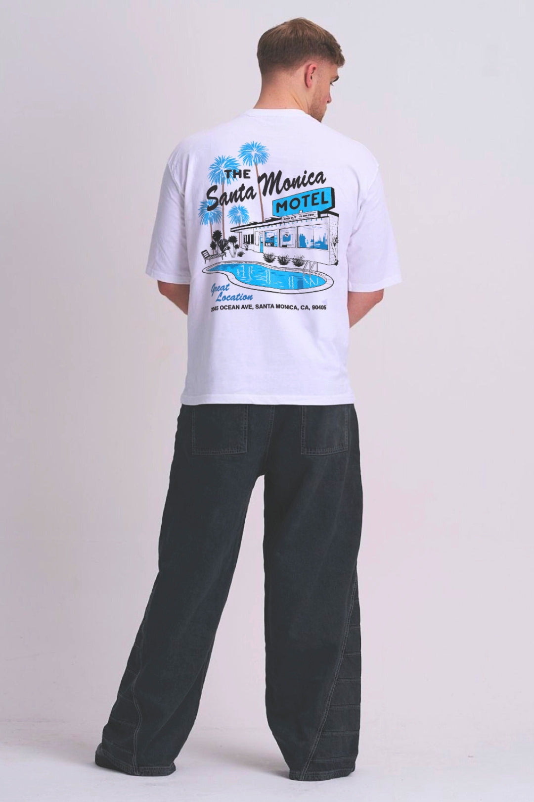 A man viewed from behind wearing a white t-shirt with a blue and black retro 'The Santa Monica Motel' graphic print, paired with dark grey wide-leg jeans, against a light grey studio background.