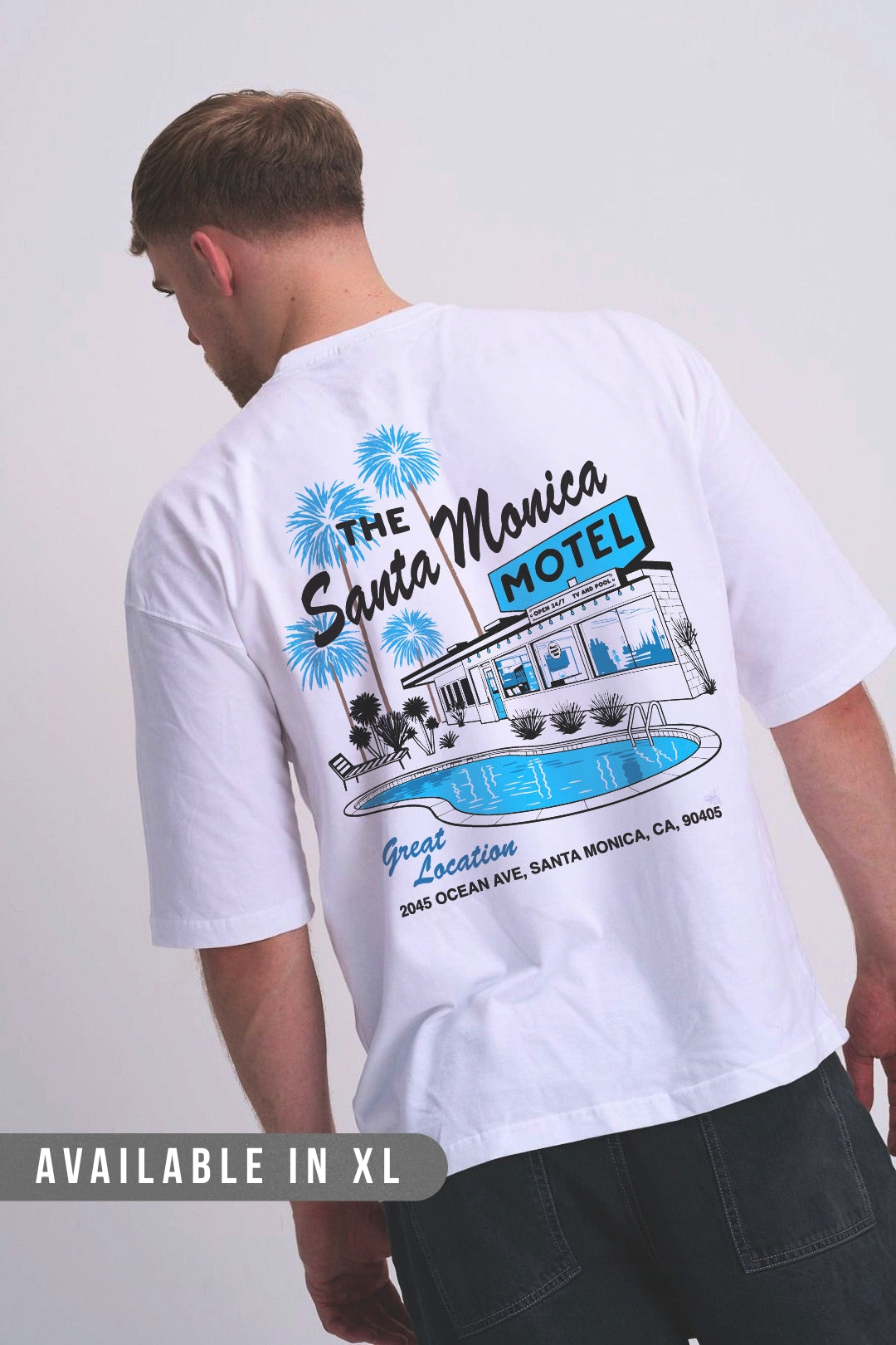 A man wearing a white t-shirt from the back, featuring a large graphic print of 'The Santa Monica Motel' with blue palm trees, a retro motel building, and a pool. 