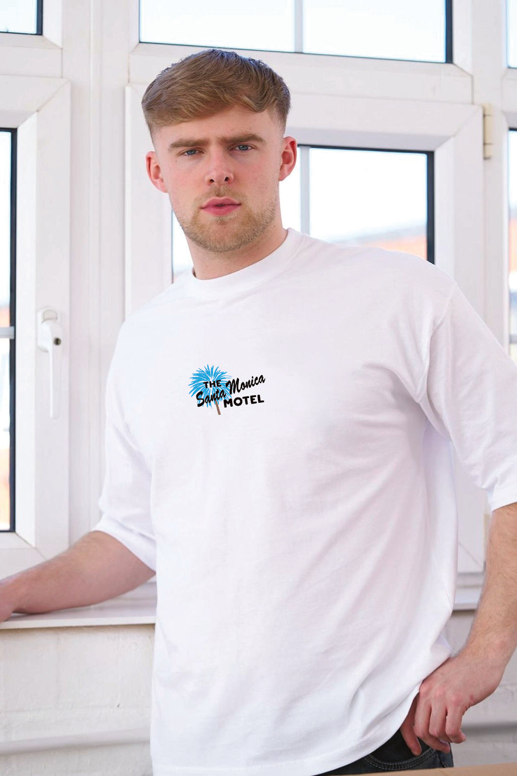 A man wearing a white short-sleeve t-shirt with a 'The Santa Monica Motel' logo printed on the left chest, standing in front of a white window frame.